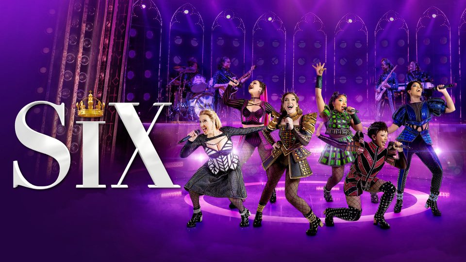 Six performers in colourful costumes sing and dance on a purple-lit stage with "SIX" text.