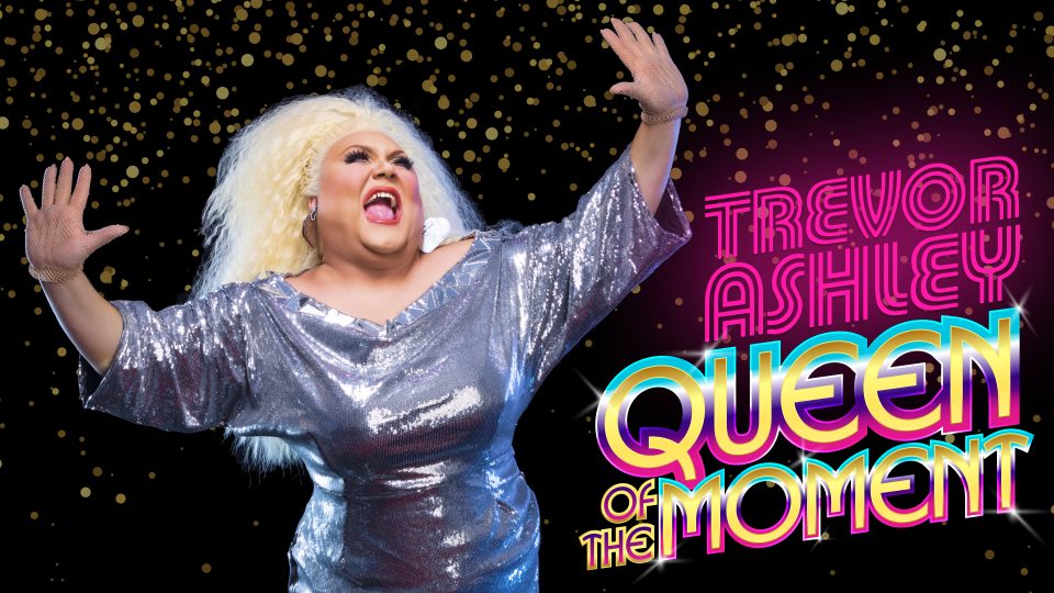 Drag performer in a silver outfit with blonde hair, posing against a sparkling background.