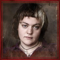 Spike Maxwell as Fate, wearing a dark suit and hat, stands against a dimly lit theatrical backdrop.