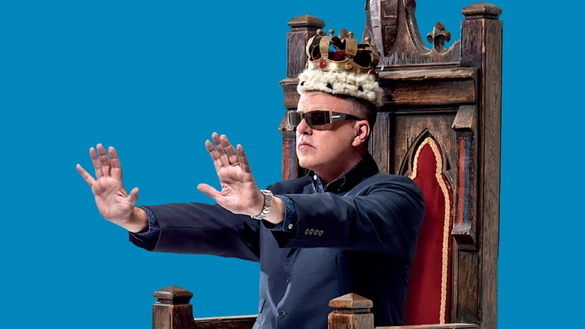 Man wearing sunglasses and a crown, seated on a throne against a blue background.