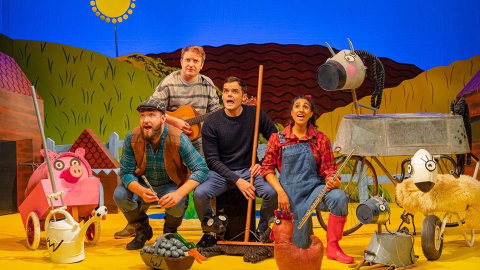 Five performers in colourful costumes, surrounded by playful farm-themed props on stage.