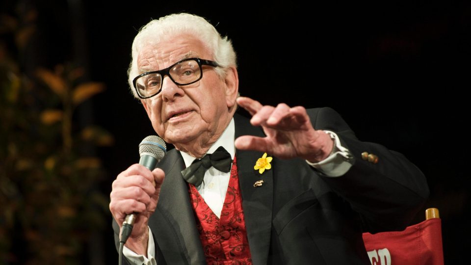 Elderly man with white hair and glasses speaking into a microphone, wearing a black suit.