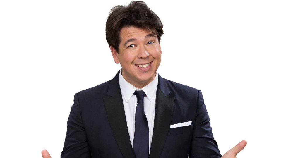 A man in a black suit smiles, gesturing with his hands against a white background.