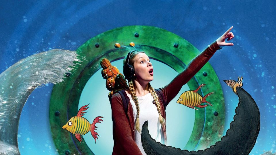 A performer with headphones points excitedly, surrounded by colourful sea creatures in a vibrant setting.