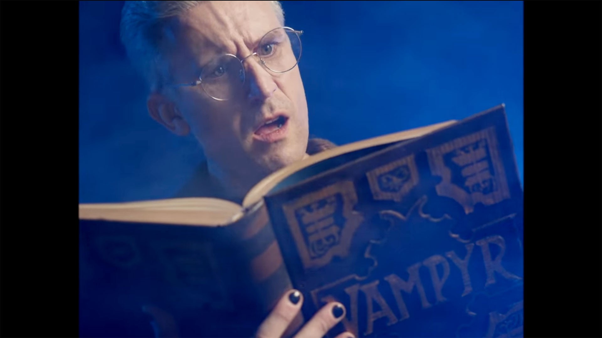 Person with glasses and painted nails reads a book titled "Vampyr" in a blue-lit setting.