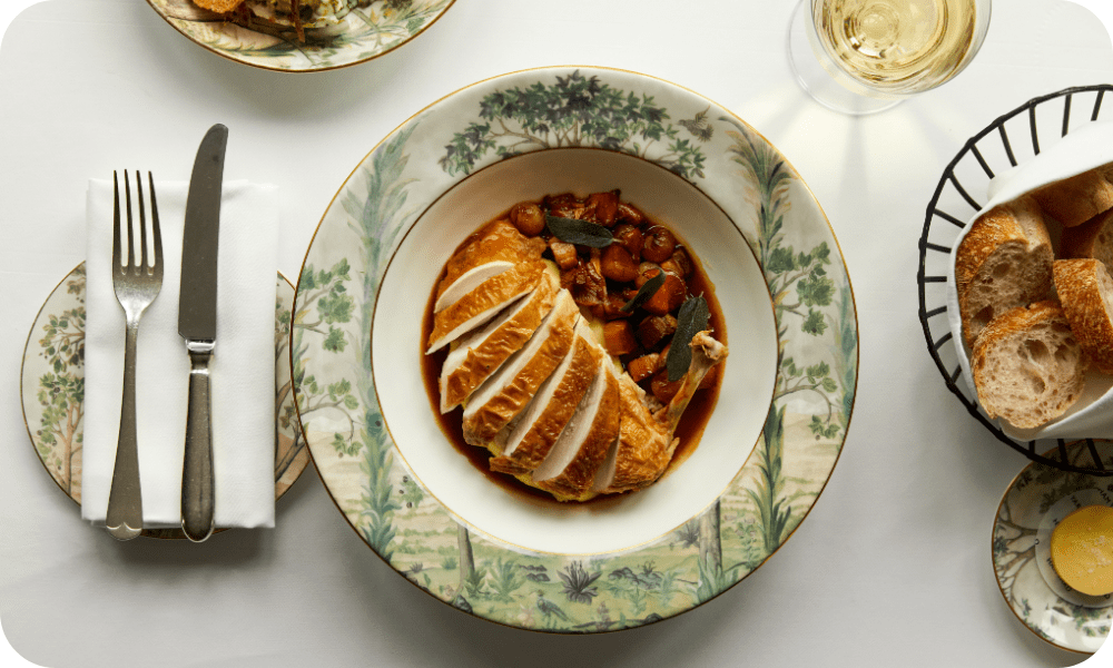 A delicious chicken dish laid out immaculately at Brasserie Max. Surrounding it is a glass of wine, bread basket, and other plates.