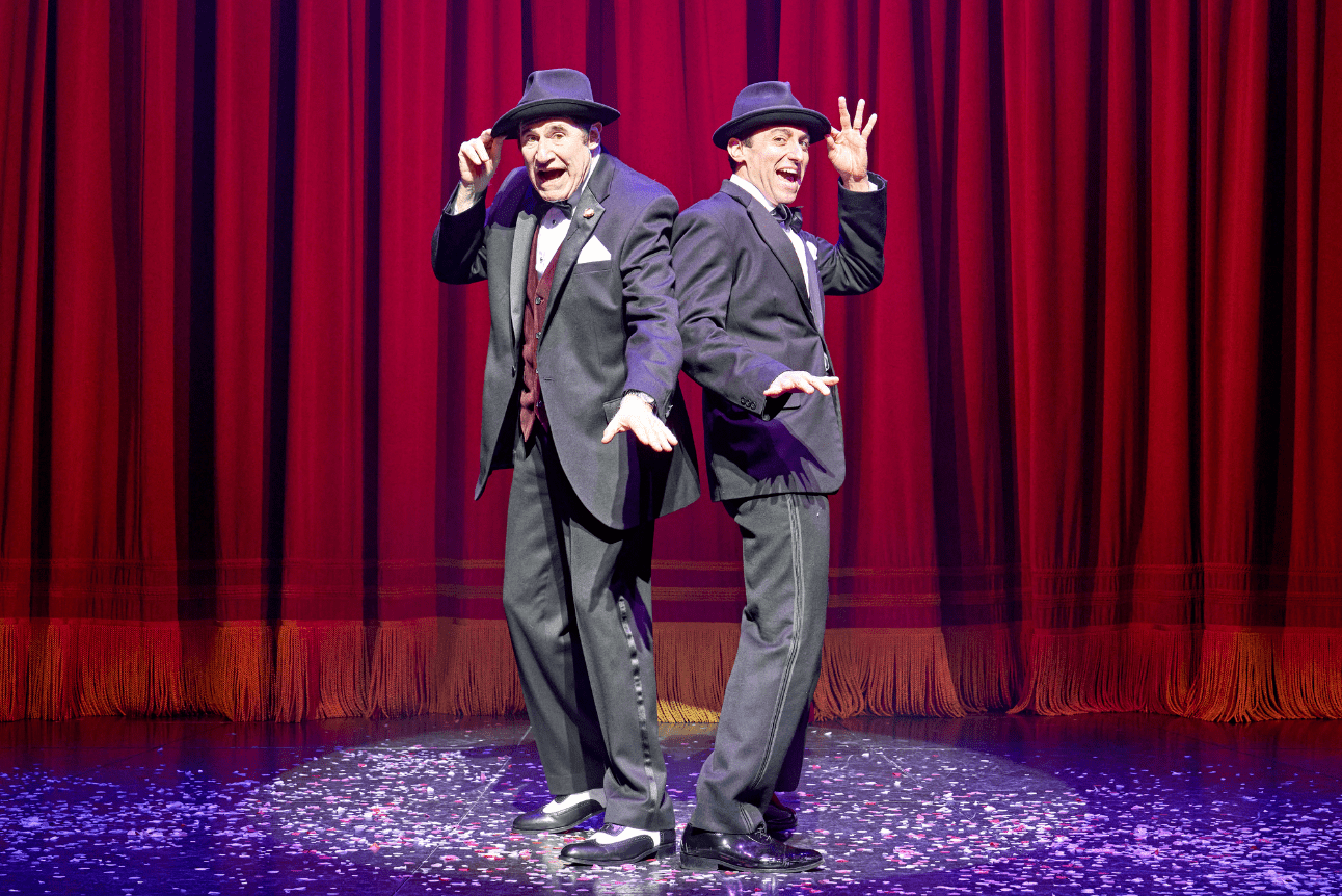 Richard Kind and Marc Antolin dance in tuxedos against a red curtain backdrop