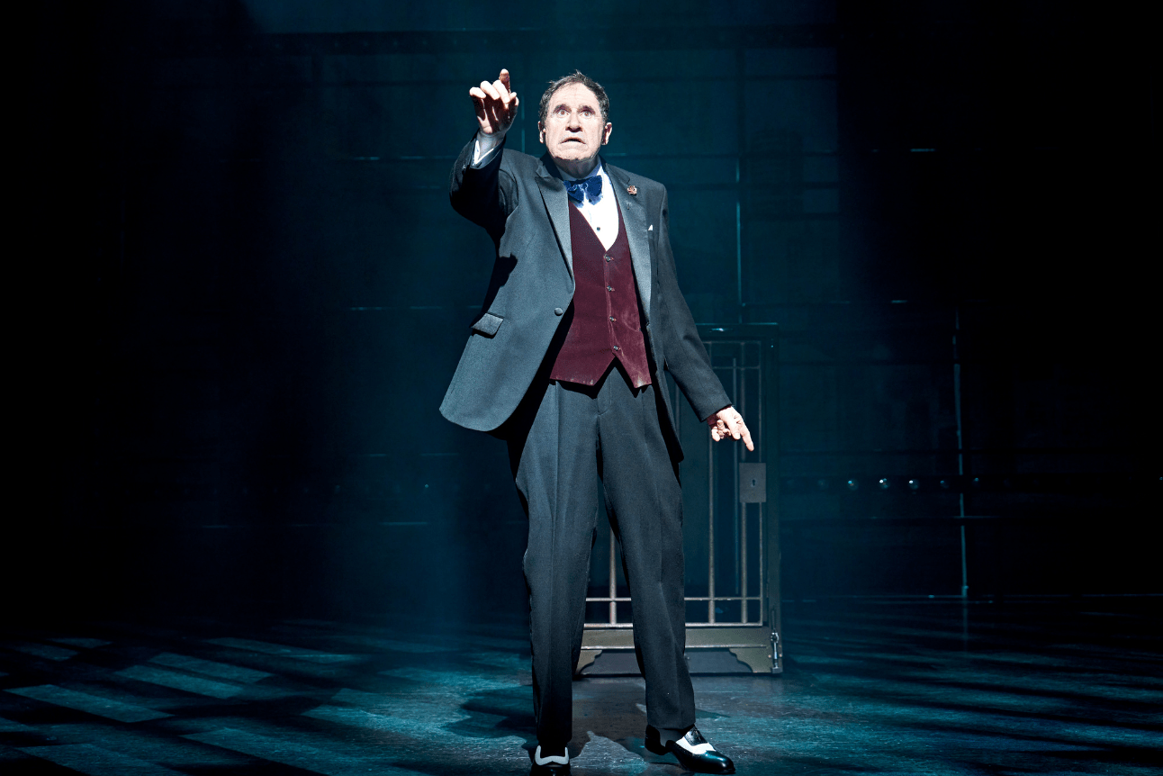 Richard Kind as Max stands with his hand held high. It's a moment of realisation.