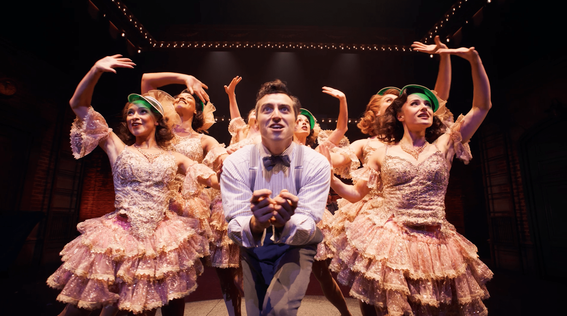A man in a striped shirt smiles, surrounded by women in pink sequined dresses and green hats.