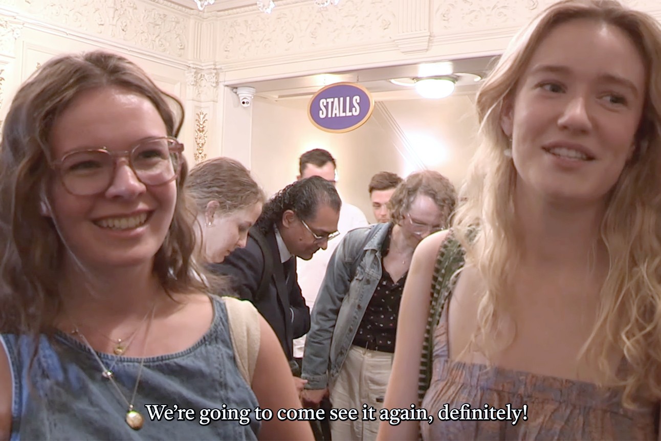 Audience members laugh and smile, seated in a dim theatre with dark walls and soft lighting.
