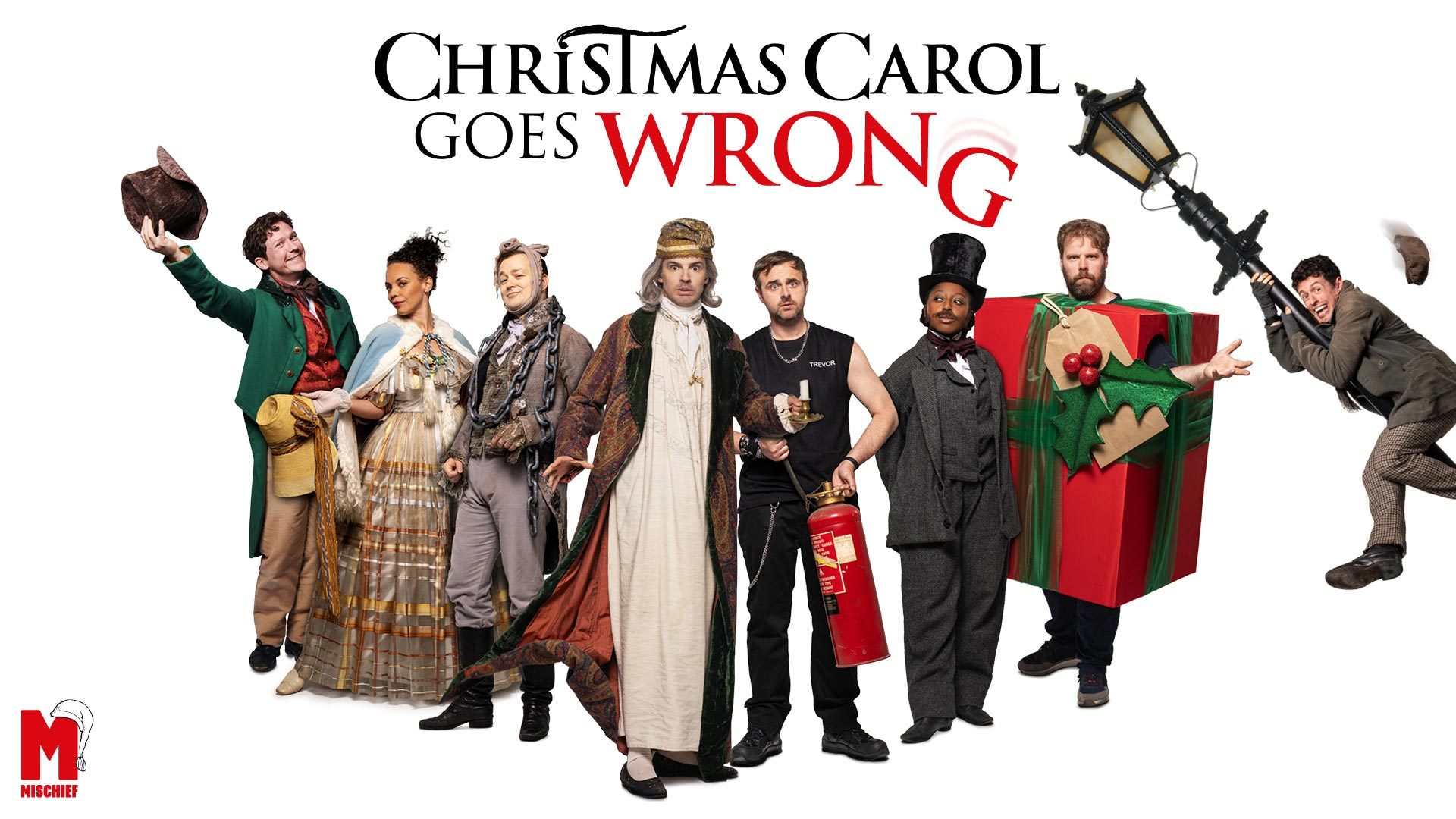 Christmas Carol Goes Wrong 1 A cast in vibrant costumes, including a large red Christmas present, poses for "Christmas Carol Goes Wrong.