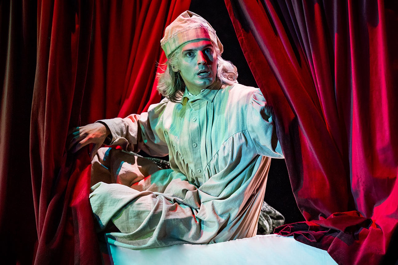 Daniel Fraser in Victorian costume, holding a candle, performing on a dimly lit stage with dark backdrop.