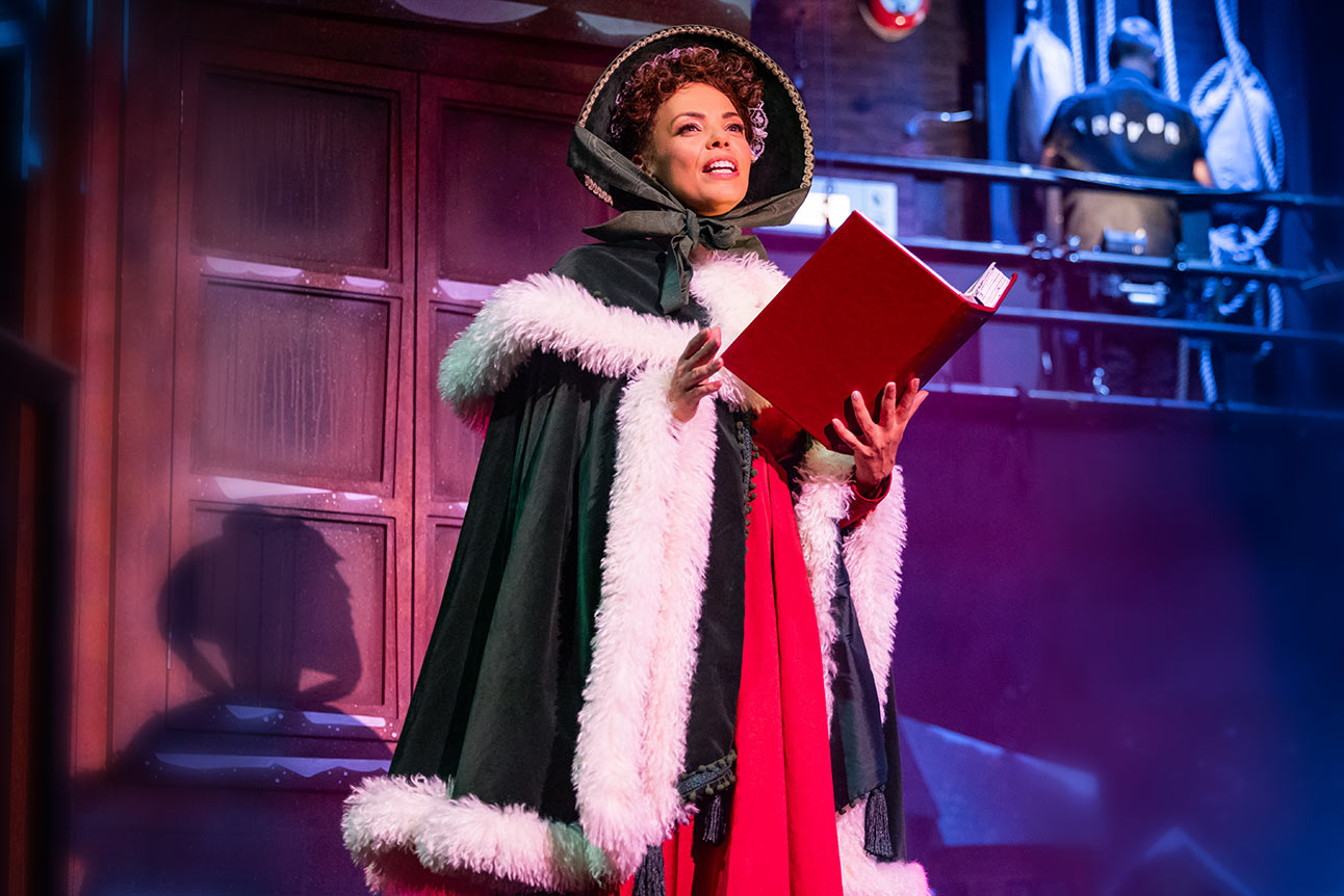 Sasha Frost smiling in festive red dress with Christmas decorations blurred in background.