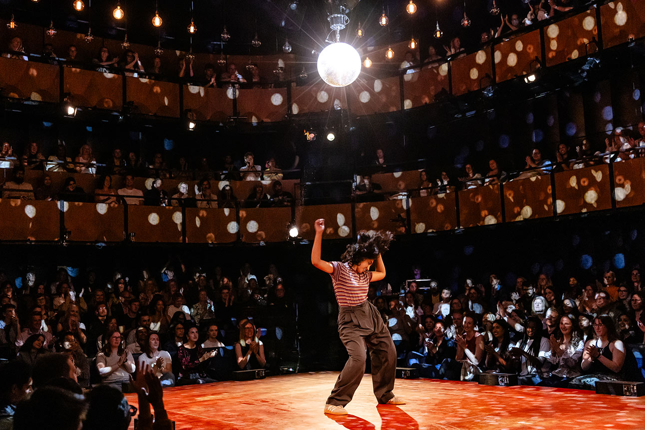 A dancer in striped attire performs on a vibrant orange stage under a sparkling disco ball.