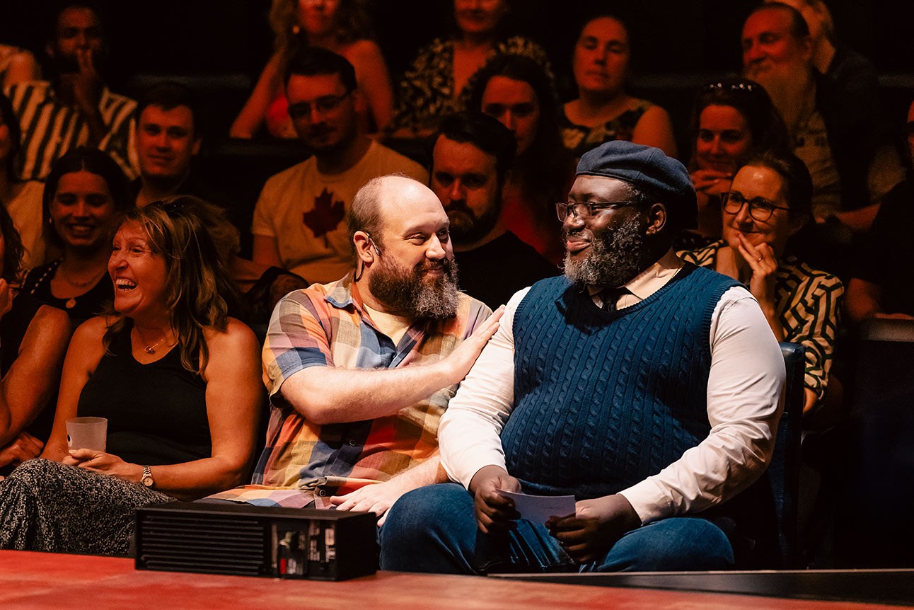 Two men share a warm interaction in a theatre, surrounded by an engaged audience in soft lighting.