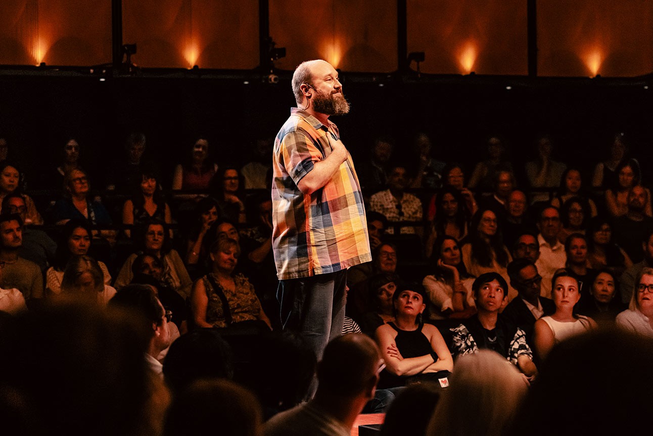 A man in a colourful plaid shirt stands on stage, addressing an engaged audience.