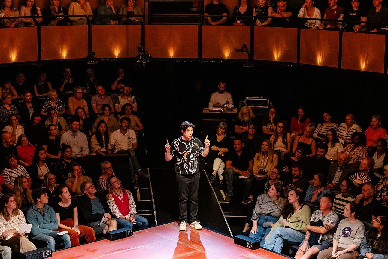 A performer in a black patterned shirt stands on a red stage, engaging with an attentive audience.