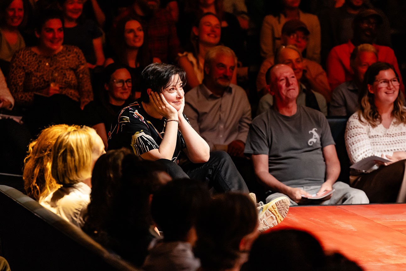 A woman with short dark hair sits thoughtfully, illuminated by warm lighting, amidst an attentive audience.