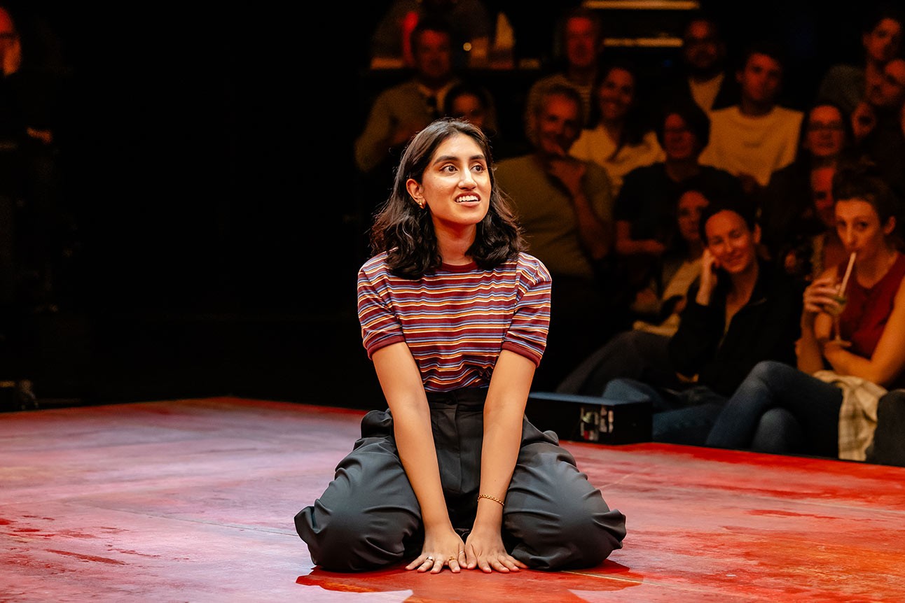 A performer in a striped top sits on a red stage, engaging with an attentive audience.