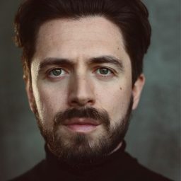 A man with dark hair and beard, wearing a black turtleneck, against a muted background.