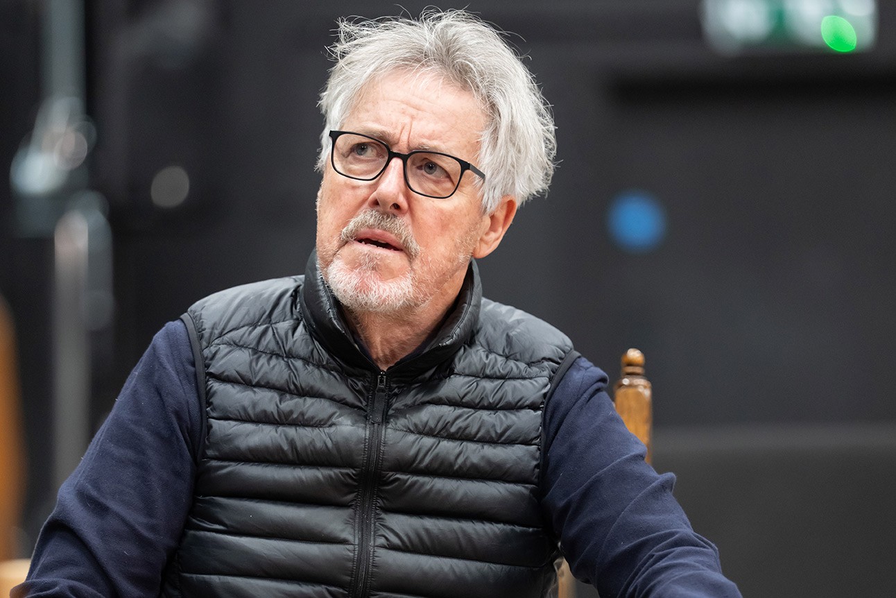 Older man with grey hair and glasses, wearing a black puffer vest and navy shirt, seated indoors.
