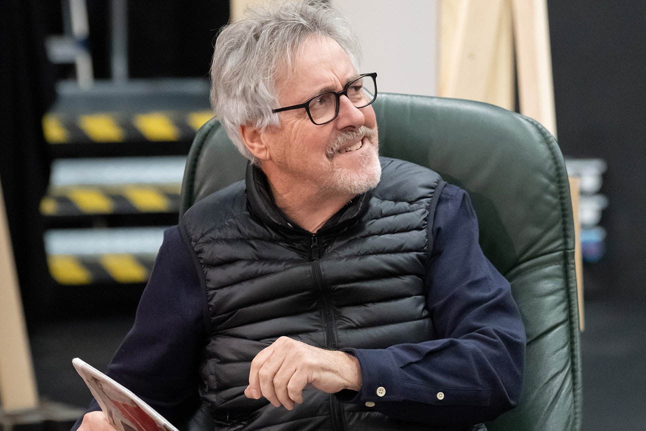 Man with grey hair and glasses wearing a black vest and navy shirt, seated indoors, smiling.