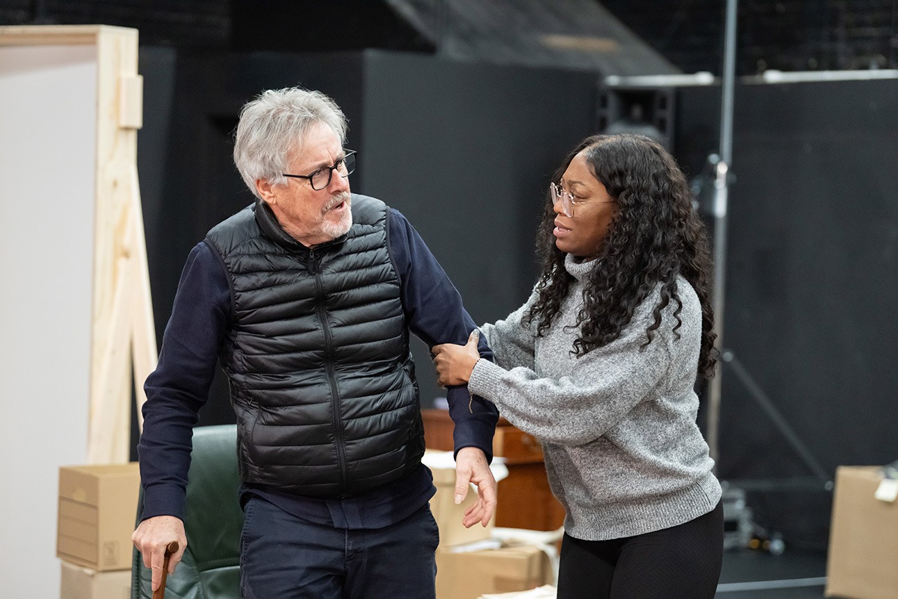 Man wearing glasses and black vest talks with woman in grey sweater, both standing in a warehouse setting.