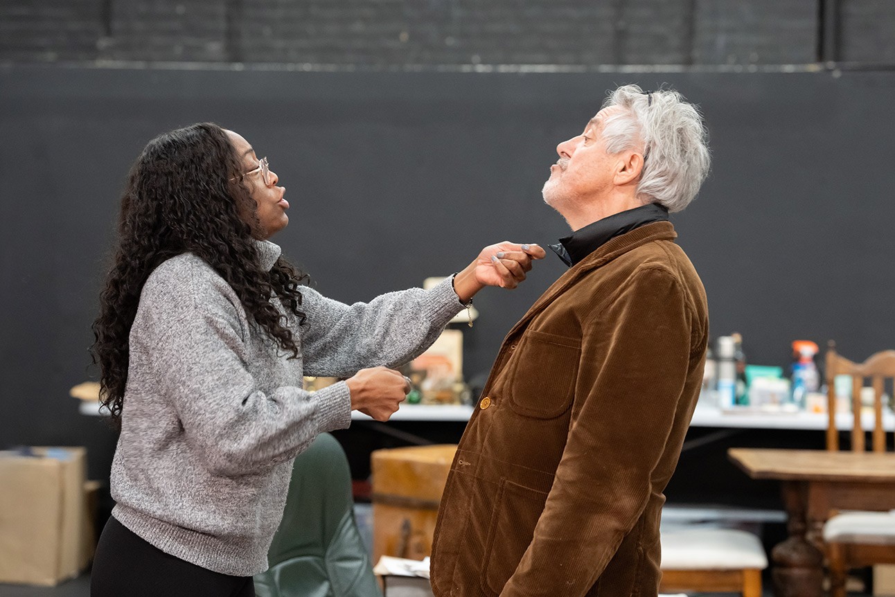 A woman adjusts a brown jacket collar of an older man with white hair against a dark background.