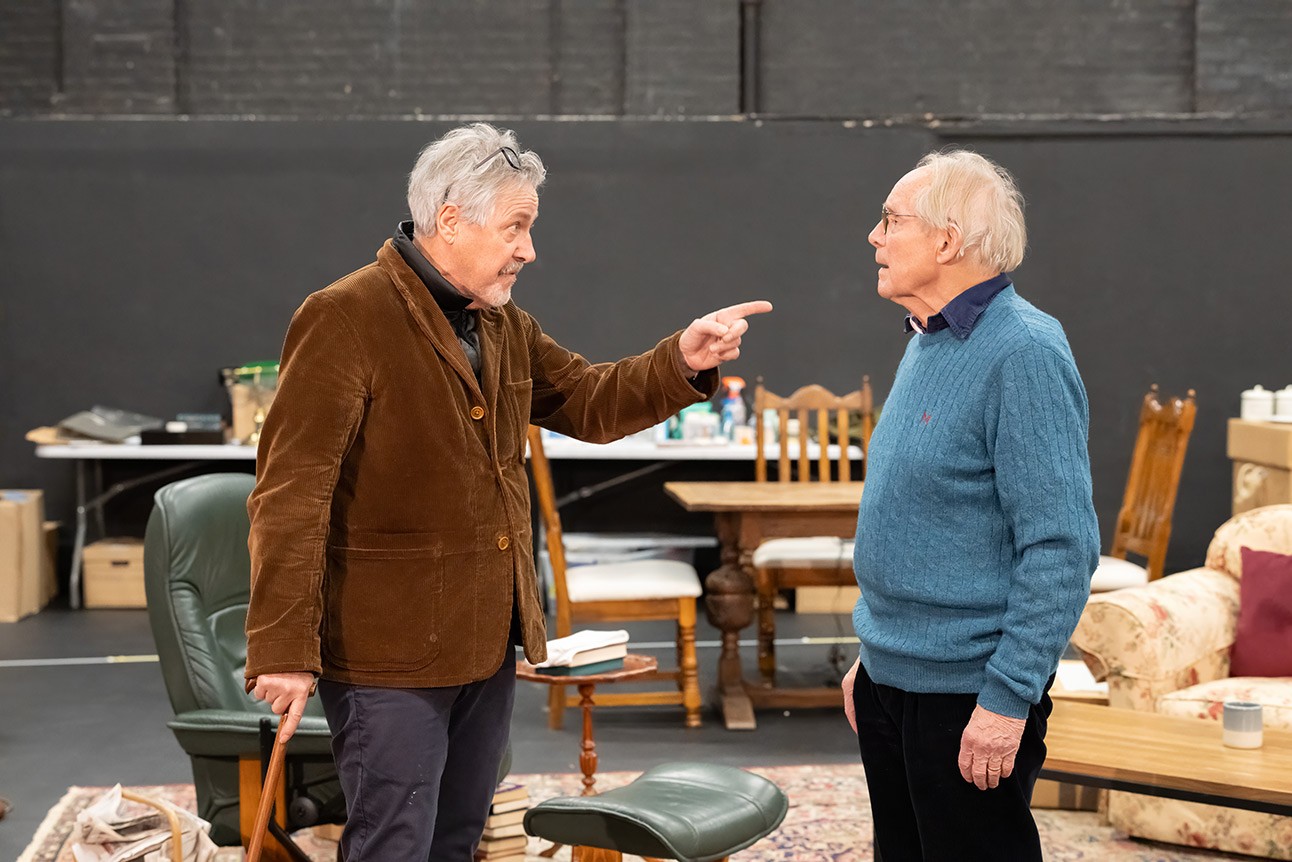 Two elderly men, one in brown jacket gesturing, the other in blue sweater listening, indoors with wooden furniture.