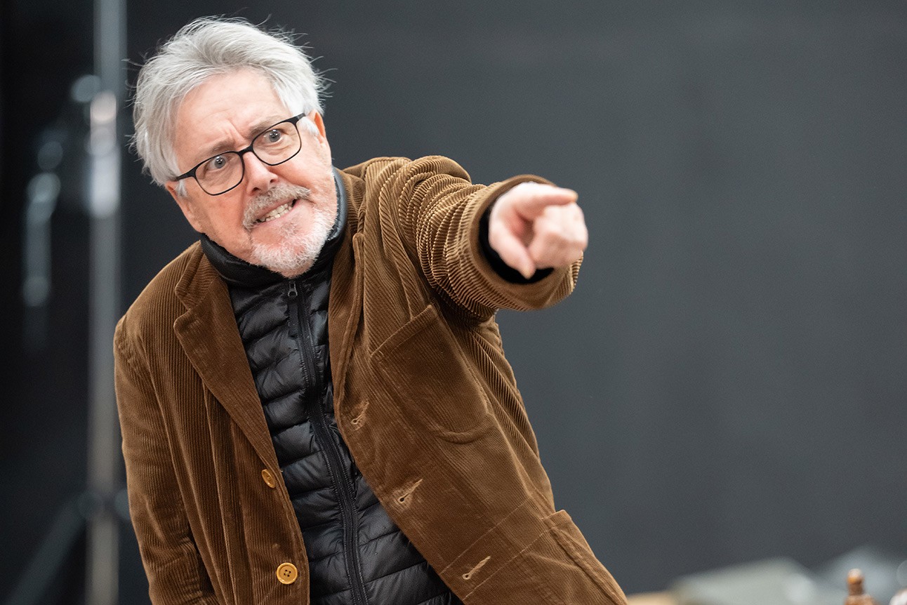 Older man with grey hair and glasses, wearing brown jacket, pointing forward against dark background.