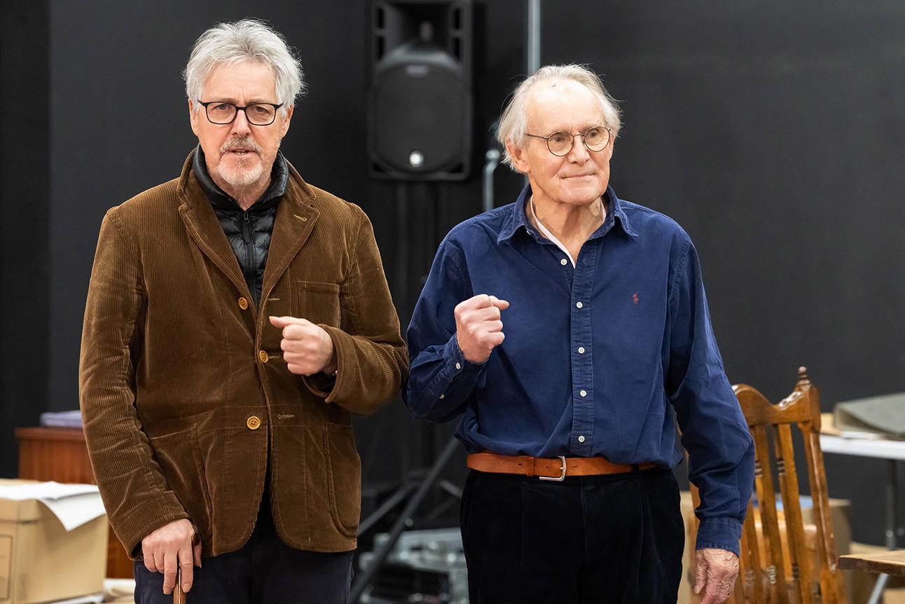 Two elderly men, one in brown jacket and glasses, the other in blue shirt, standing against dark background.