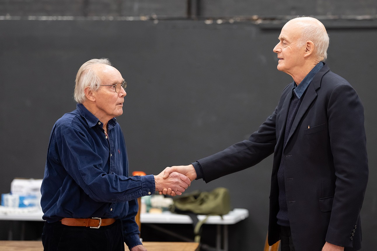 Prime Minister and older man shake hands indoors, dark background, both wearing dark jackets.