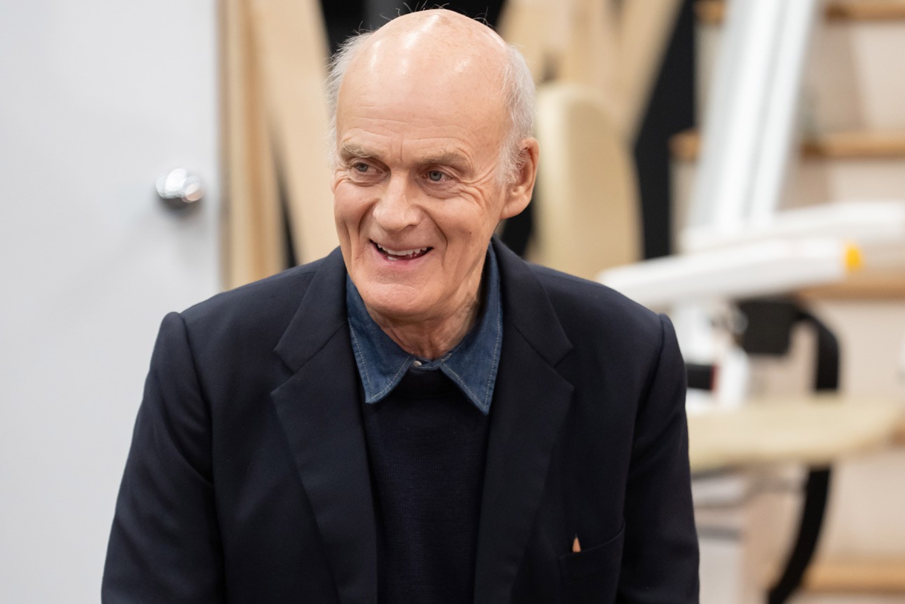 Elderly man with a bald head, wearing a dark jacket and blue shirt, smiling indoors.