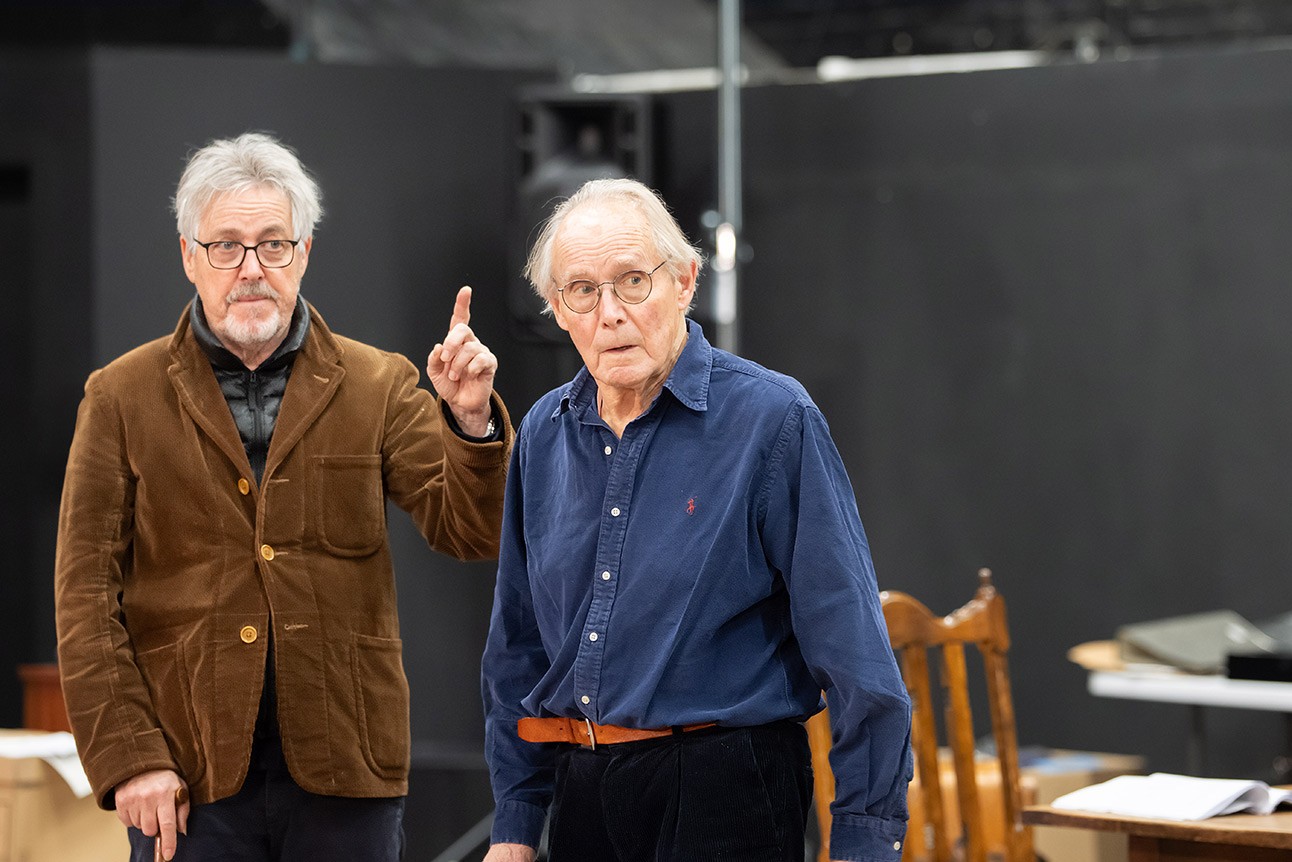 Two older men, one in brown jacket pointing, the other in blue shirt gesturing, black background.