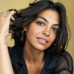 A person with dark hair and hoop earrings touches their hair, wearing a black shirt, yellow background.