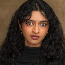 A person with long curly hair and a black top against a brown textured background.