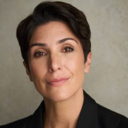 A woman with short dark hair, wearing a black blazer, poses against a neutral background.