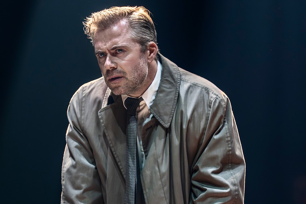Man with slicked-back hair wearing a beige trench coat and dark tie against a dark background.