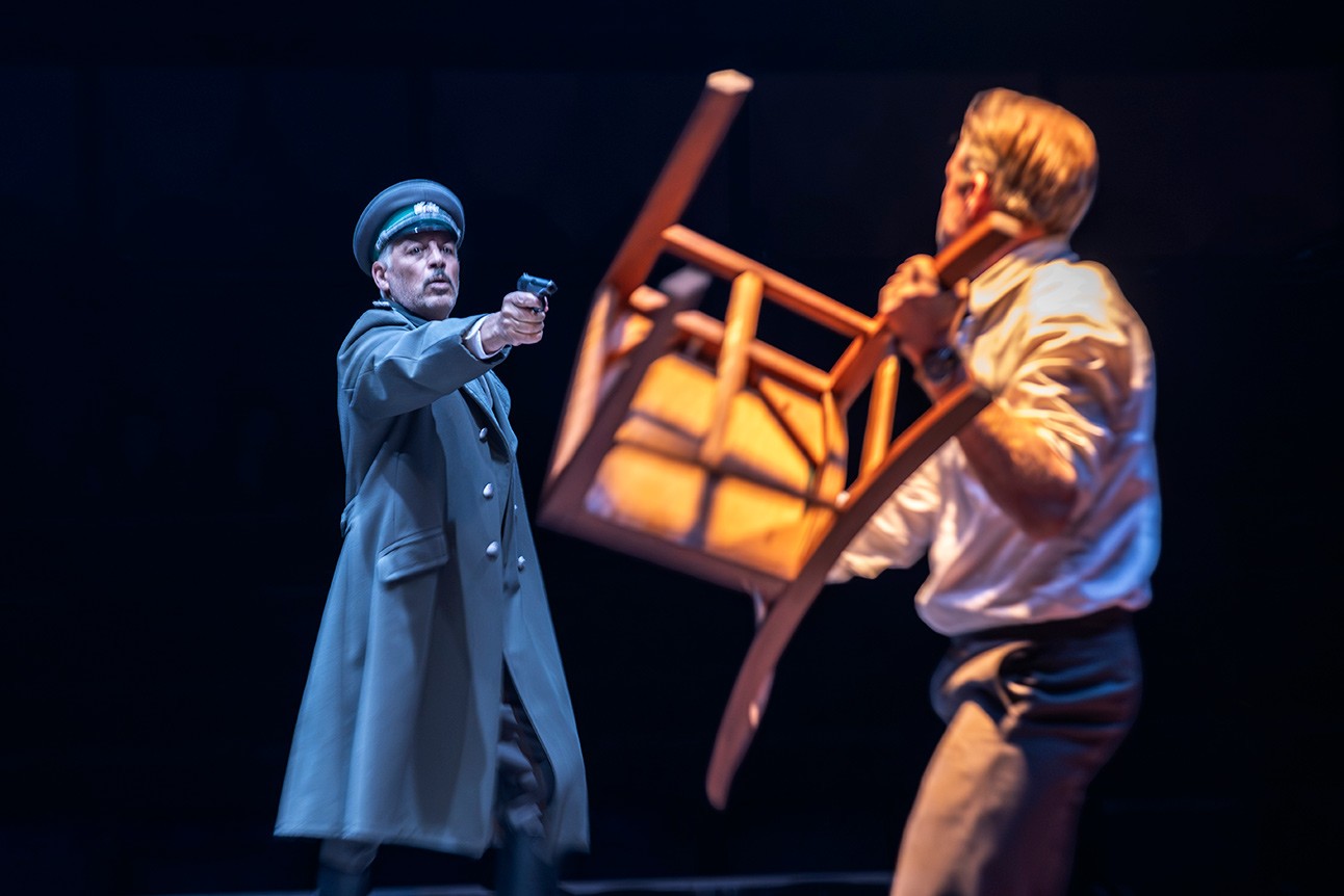 Actor in dark coat and hat points gun at another actor holding a chair, on stage with dark background.