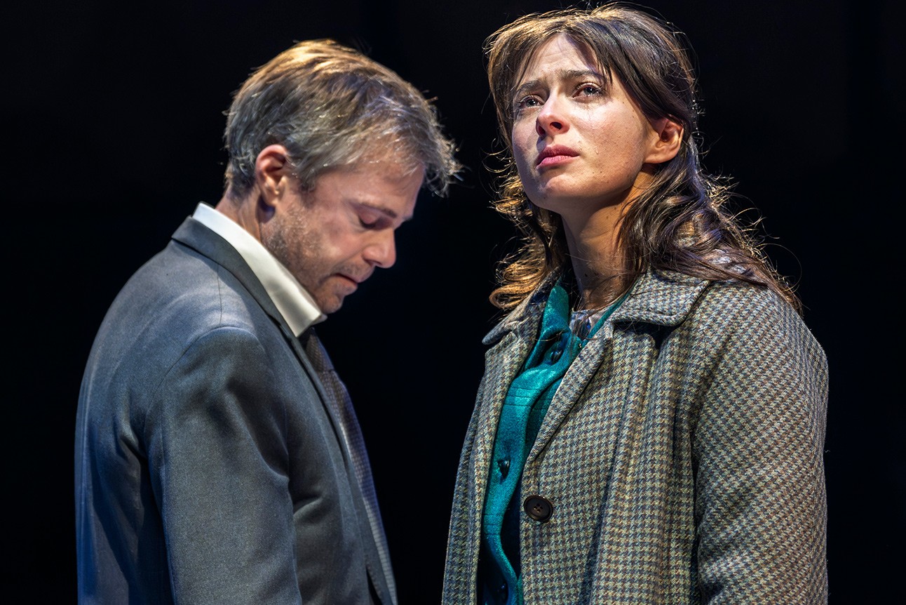 Two actors in serious expressions; man in dark suit looks down, woman in checked coat gazes upward.