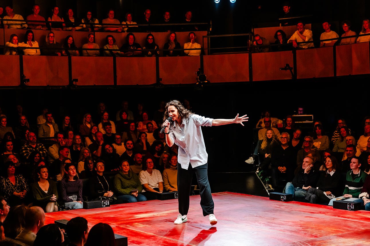 A performer in a light shirt gestures passionately on a red stage, with an engaged audience behind.
