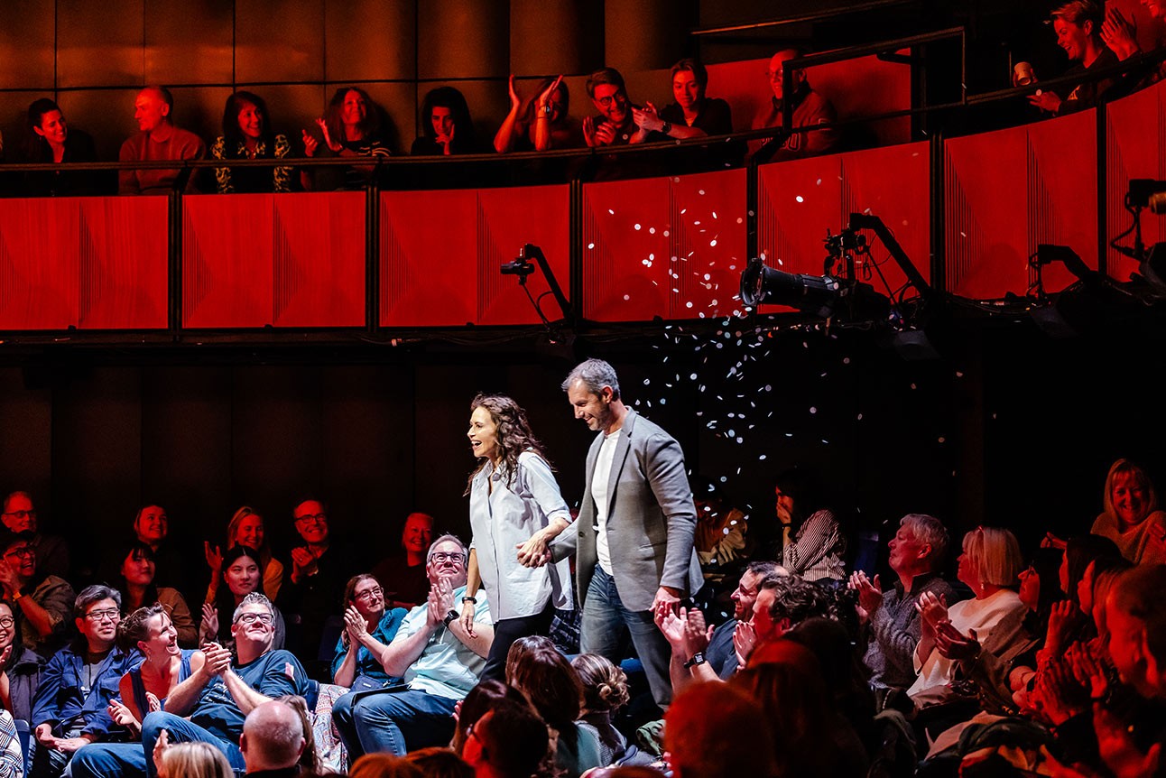 A woman in a light shirt and a man in a grey blazer walk hand-in-hand through a cheering audience.
