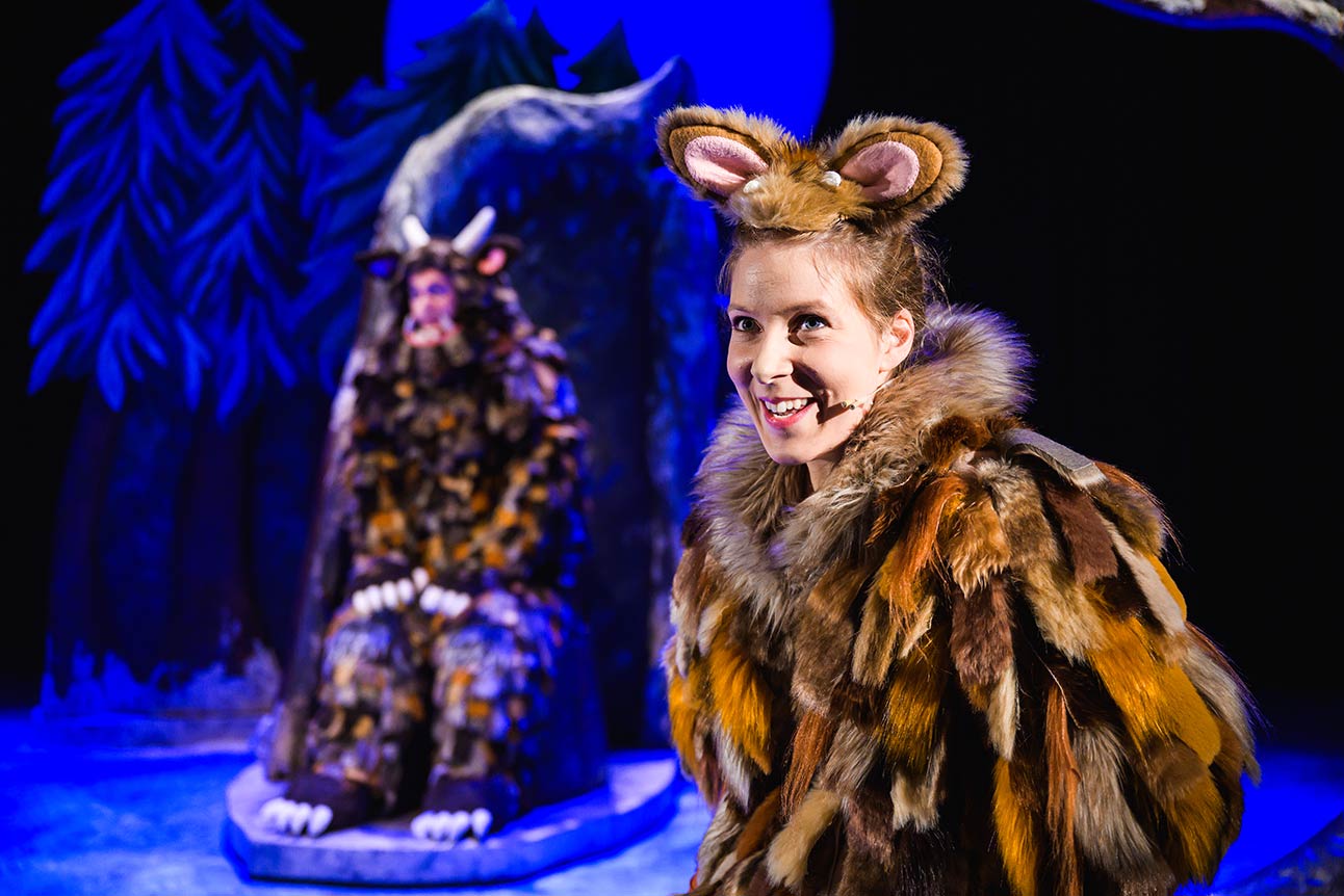 A performer in a brown and orange fur costume smiles, with a seated figure in the background.