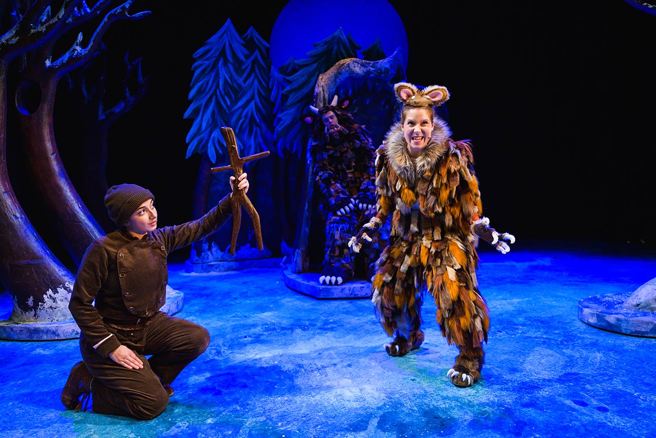 A performer in a furry brown costume grins, while another kneels with a stick on a blue stage.