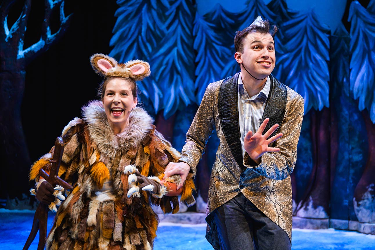 Actors in vibrant costumes perform on stage, surrounded by blue and green forest-themed scenery.