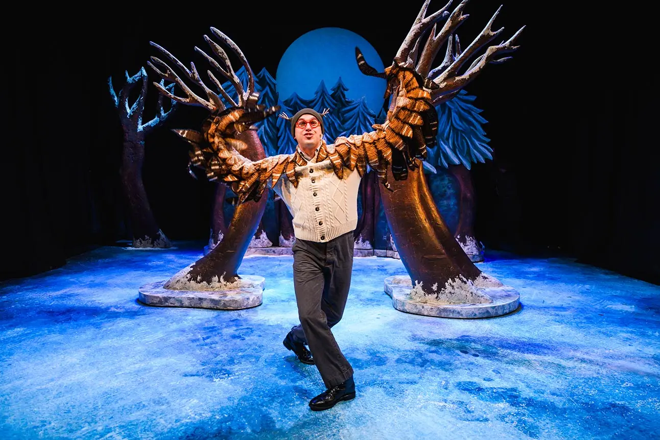 A performer in a cream sweater poses dramatically against a blue, icy stage backdrop with trees.