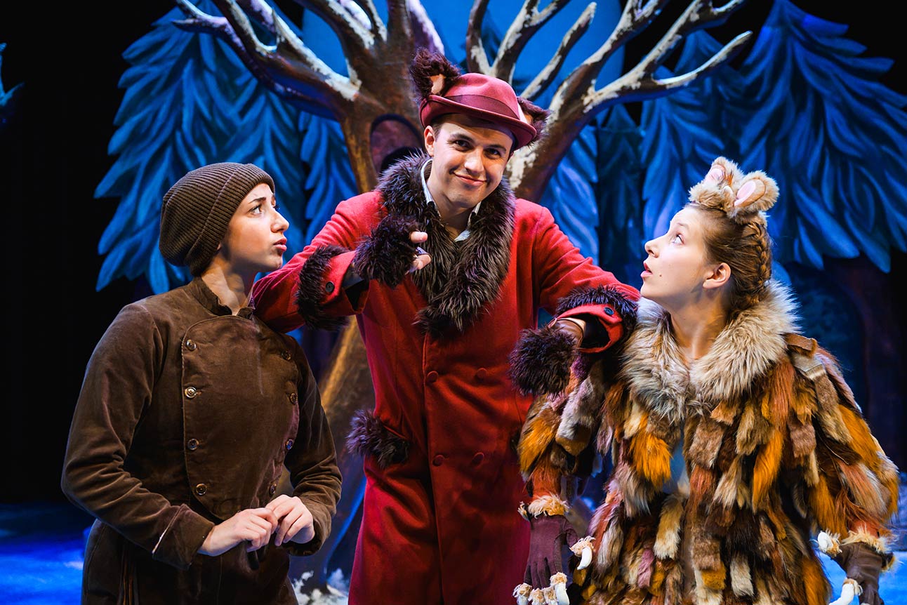 Three actors in textured costumes interact on stage, with vibrant blue trees and a dark backdrop.