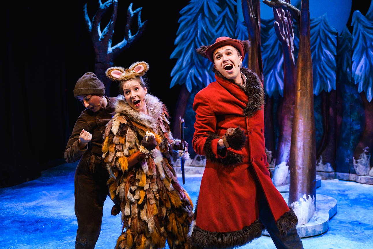 Three performers in animal costumes, including a red coat, pose joyfully on a blue stage.
