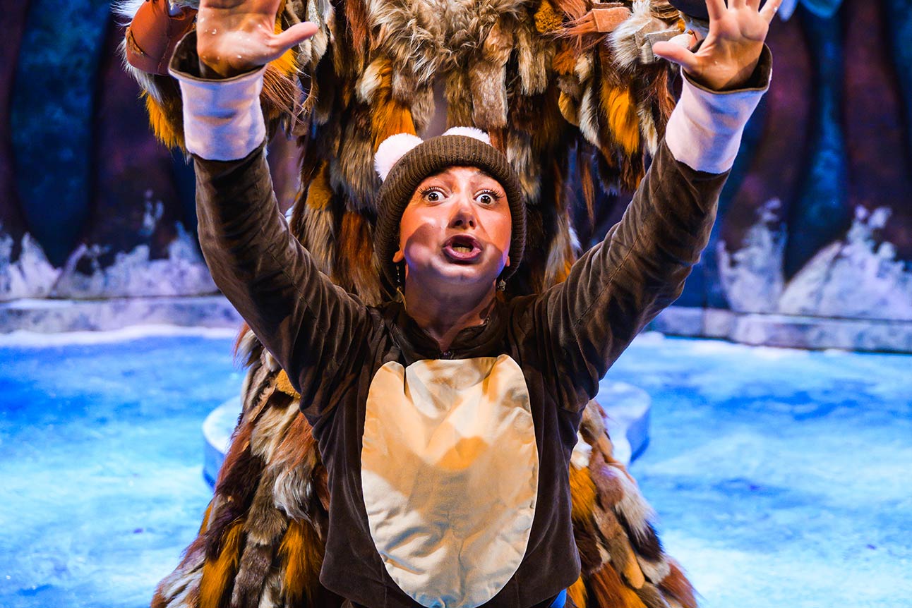 A performer in a brown costume with a white belly gestures dramatically against a snowy backdrop.