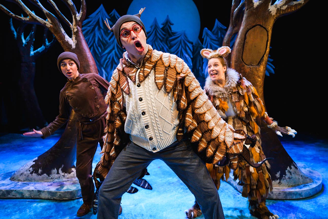 Three actors in whimsical costumes perform on a blue stage with stylised trees and a moon backdrop.